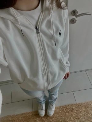 Vit zip-hoodie från Ralph Lauren(ny) - Säljer en fräsch vit hoodie med dragkedja från Ralph Lauren. Hoodien har huva med snören, två fickor framtill och den klassiska broderade loggan på bröstet. Perfekt för en chill och stilren look.