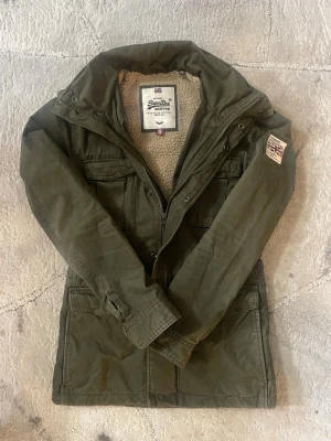 Superdry Military Field-jacket - Säljer en snygg och varm Superdry Military-jacka i mörk olivgrön färg, storlek S. Jackan har mjukt teddyfoder på insidan, flera praktiska fickor och en militärinspirerad design. Perfekt för höst och vinter.  💬 pris kan diskuteras vid snabbt köp
