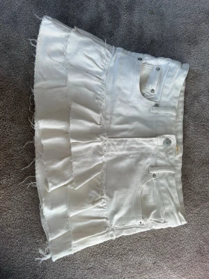 Själv gjord vit jeanskjol med volangdetalj  - Vit kort jeanskjol med volangdetalj och råa kanter. Kjolen har klassiska fickor fram och bak samt knapp- och dragkedjestängning. Perfekt för dig som gillar en trendig och avslappnad stil.