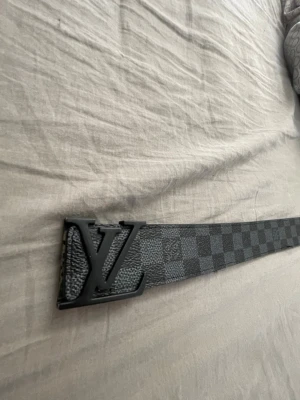 Svart rutigt  bälte - Snyggt svart bälte med rutigt mönster och stor logga på metallspännet. Bältet har en klassisk design och passar perfekt till jeans eller kostymbyxor. Insidan är beige och utsidan har en matt finish.lite använd 