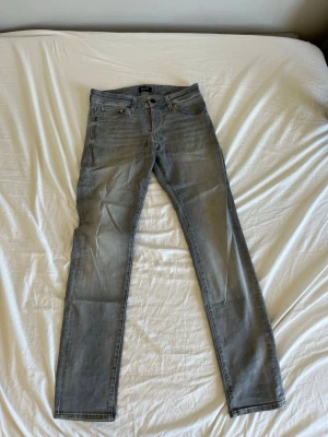 Gråa raka jeans - Säljer ett par gråa jeans med rak passform och klassisk femficksdesign. Jeansen har normal midja och dragkedjegylf. Materialet är bomullsdenim med lite stretch för extra komfort. Perfekta till en avslappnad stil.