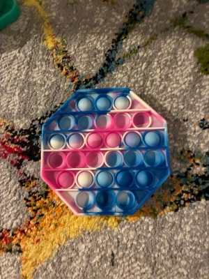Flerfärgad pop it i silikon - Säljer en åttakantig pop it i silikon med blå, rosa och vita fält. Perfekt för att stressa ner eller bara ha något att pilla med. Lätt att ta med sig och enkel att använda, tryck på bubblorna och få ett tillfredsställande popp-ljud.