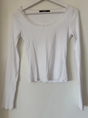 Vit långärmad topp från BikBok - Säljer en söt vit långärmad topp från BikBok. Toppen är ribbad och har spetsdetaljer runt den runda halsringningen samt en liten rosett framtill. Perfekt att styla med jeans eller kjol för en enkel men gullig look.