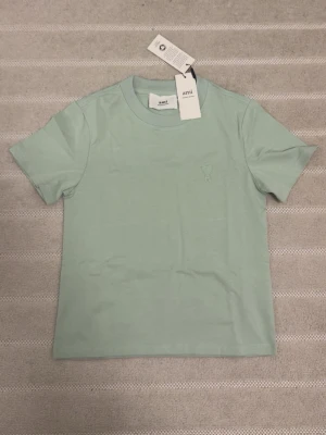 Mintgrön t-shirt från Ami Paris - Fräsch mintgrön t-shirt från Ami Paris med diskret broderad logga på bröstet. Klassisk rund hals och korta ärmar. Tillverkad i mjuk bomull för en skön känsla. Perfekt till jeans eller shorts för en clean look. Hade passat ett barn runt 5 års åldern.