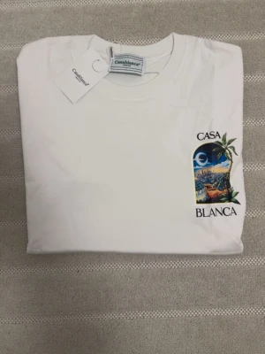 Vit t-shirt från Casablanca - Vit t-shirt från Casablanca med tryck på bröstet som visar ett färgstarkt motiv med palmer och texten 'Casa Blanca'. Klassisk rund hals och korta ärmar. Perfekt för en avslappnad och stilren look.
