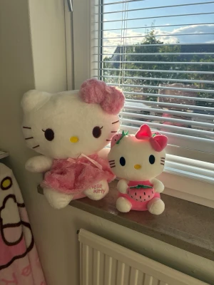 Två Hello Kitty gosedjur rosa tema - Säljer två söta Hello Kitty gosedjur. Ena har en fluffig rosa klänning och stor rosett, den andra har en jordgubbsdräkt med rosa rosett och gröna detaljer. Perfekta som dekoration eller samlarpryl för Hello Kitty-fans. Går att köpa varförsig I Dm. Ca 35x30cm och 25x20cm