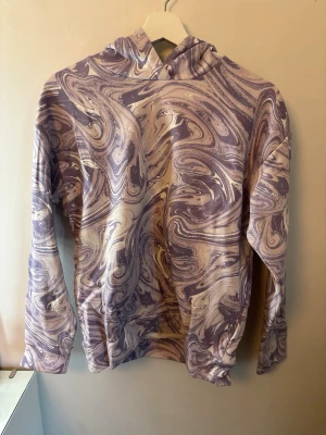 Lila och vit swirl hoodie - Säljer en hoodie med unikt swirl-mönster i lila och vitt. Hoodien har huva och långa ärmar, samt en avslappnad passform. Perfekt för dig som gillar färgglada och kreativa plagg. Materialet känns mjukt och bekvämt mot huden.