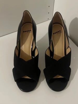 Svarta pumps med öppen tå  - Snygga svarta pumps med öppen tå och korslagda band framtill. Skorna har en elegant klack och är tillverkade i en kombination av mocka och läder. Perfekta för dig som vill ha en stilren och trendig look.