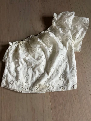 Vit offshoulder blus med broderi - Superfin vit offshoulder blus med volang och broderade blommor. Toppen har en ärm och är gjord i bomull, perfekt för en somrig look. Söt och luftig modell med romantiska detaljer.