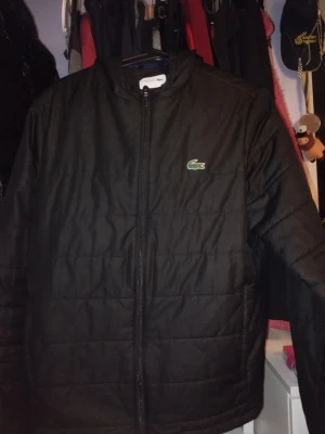 Lacoste Sport-jacka med lätt vaddering - Svart pufferjacka från Lacoste med klassisk grön krokodillogga på bröstet. Jackan har dragkedja framtill och blått foder på insidan. Perfekt för kalla dagar och har en stilren, enkel design som funkar till det mesta.
