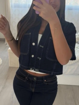 Kort mörkblå jeansjacka - Snygg kort jeansjacka i mörkblå denim med kontrastsömmar och stora framfickor. Jackan har korta ärmar, silverfärgade knappar och en boxy fit som ger en trendig look. Perfekt att styla med höga jeans eller kjol för en cool vibe.