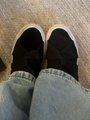Svarta slip-on sneakers med knut - Säljer ett par svarta slip-on sneakers med breda tygband i knut över foten och vit sula. Storlek 36. endast använda några få gånger. 