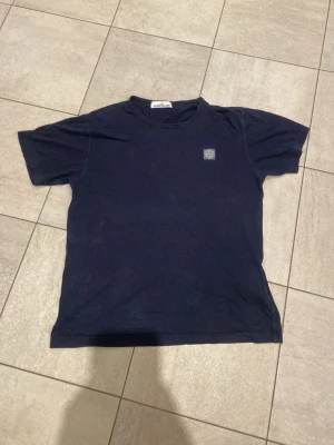 Mörkblå Stone Island t-shirt - Säljer en mörkblå t-shirt från Stone Island med klassisk logopatch på bröstet. T-shirten har rund hals och korta ärmar, perfekt för en clean och stilren look. Materialet känns mjukt och bekvämt mot huden. Lite lös runt nacken.