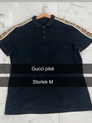 Svart Gucci pikétröja - Snygg svart pikétröja från Gucci med klassisk krage och knappslå. Ärmarna har beige band med bruna Gucci-loggor längs axlarna för en exklusiv look. Perfekt för dig som vill ha en stilren men ändå lyxig topp.