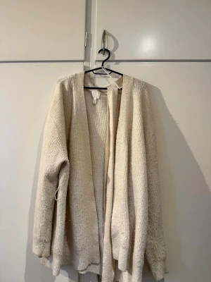 Beige lång kofta från H&M - Mysig beige lång kofta från H&M med öppen front och ribbad struktur. Koftan har långa ärmar och ett avslappnat, oversized snitt som passar perfekt för lager på lager. Tillverkad i mjukt stickat material som känns skönt mot huden.