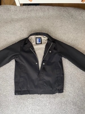 Jack & Jones svart Grisch jacka  - Snygg Grisch jacka  från Jack and Jones med rutigt innerfoder i beige och vitt. Jackan har klassisk krage, dragkedja och tryckknappar framtill samt två sidofickor. Storlek = 152cm men passar även 158cm. 