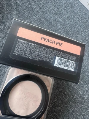 Huda Beauty Easy Bake Peach Pie - Foundation i löspuderformat från Huda Beauty i nyansen Peach Pie. Kommer i en fyrkantig burk med svart lock och transparent botten. Pudret har en ljus, persikotonad färg som ger en naturlig finish.