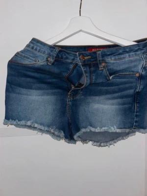Blå jeansshorts med fransig kant - Säljer ett par klassiska blå jeansshorts med fransig, rå kant och femficksmodell. Shortsen har normal passform och är perfekta för varma dagar. De har en snygg tvättad look och stängs med knapp och dragkedja.