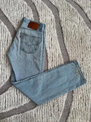 Levi's 501 ljusblå raka jeans - Klassiska Levi's 501 jeans i ljusblå denim med rak passform och hög midja. Jeansen har fem fickor, knappgylf och den ikoniska läderpatchen bak i midjan. Perfekta för en avslappnad och tidlös stil.