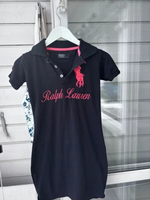 Svart kort pikéklänning Ralph Lauren - Svart pikéklänning från Ralph Lauren med rosa broderad logga och text framtill. Klänningen har korta ärmar, krage och knappar vid halsen. Rosa detalj med siffran 2 på ena ärmen. Perfekt för en sportig och avslappnad look.