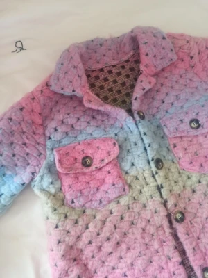 Pastellfärgad stickad jacka - Unik stickad jacka i pastellfärger med rosa, blå och lila toner. Jackan har två stora bröstfickor med knappar, krage och knäppning framtill. Grovstickad struktur och mjukt material, perfekt för dig som gillar färg och retrostil.