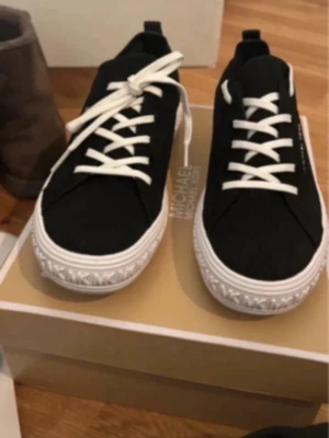 Svarta sneakers med vit sula - Stilrena svarta sneakers från Michael Kors med vit, räfflad sula och vita snören. Skorna har en låg profil och diskret logga på sidan. Perfekta för dig som gillar enkel men snygg design. Materialet ser ut att vara canvas eller textil.