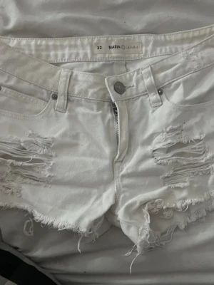 Vita slitna jeansshorts från Maria Denim - Vita jeansshorts från Maria Denim med snygga slitningar och fransig kant. Klassisk femficksmodell med dragkedja och knapp framtill. Perfekta för en avslappnad och trendig sommarstil.