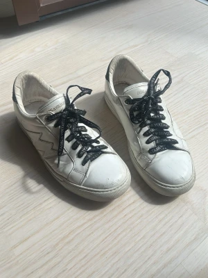 Zadig&Voltaire Sneakers - Snygga vita sneakers med en blixtdetalj på sidan och svarta skosnören med Zadig & Voltaire text. Skorna har en låg profil och vit sula. Materialet är slätt läder och hälen har en svart glitter+kontrastdetalj. Perfekta för en cool och avslappnad stil.