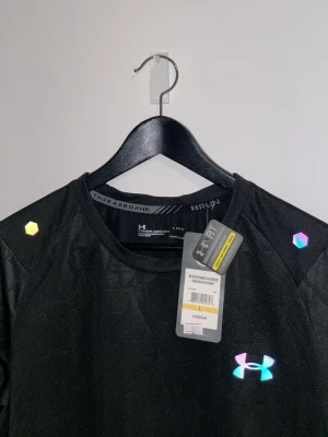 Svart tränings-tshirt från Under Armour - Svart t-shirt från Under Armour med rund hals och reflekterande logga framtill. Tillverkad i ett lätt och tekniskt material som andas, perfekt för löpning eller träning. Diskreta reflexdetaljer på axlar och rygg för extra synlighet.
