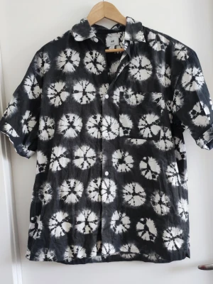 Svart och vit batikskjorta från H&M - Säljer en kortärmad skjorta från H&M med svart och vit batikmönster. Skjortan har klassisk krage, bröstficka och knäppning framtill. Perfekt för dig som gillar en avslappnad och trendig stil.