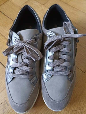 Grå sneakers med mockadetaljer - Snygga grå sneakers med både mocka och glansiga paneler, vita sulor och matchande grå skosnören. Skorna har dragkedja på sidan och coola detaljer som ger en modern look. Perfekta för dig som gillar stilrena och neutrala färger.