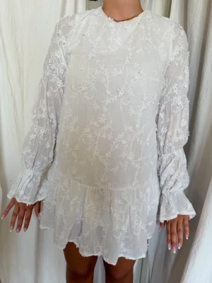 Vit blommig kort klänning med volang - Säljer en söt vit kort klänning med blommigt broderat mönster. Klänningen har långärmade, lätt puffiga ärmar med volang vid ärmslut och nederkant. Tillverkad i ett tunt, luftigt material som passar perfekt till sommaren.