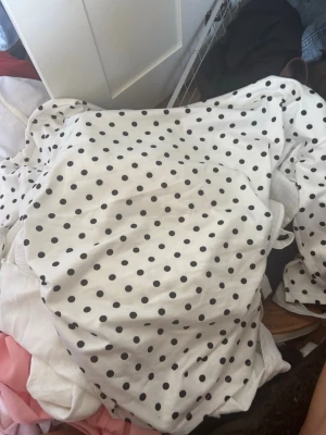Vit prickig kortklänning med rynk - Säljer en vit kortklänning med svarta prickar och snygga rynkdetaljer på sidan. Klänningen har en klassisk och lekfull look, perfekt för dig som gillar mönster. Materialet känns mjukt och lätt, troligen bomull eller polyester.