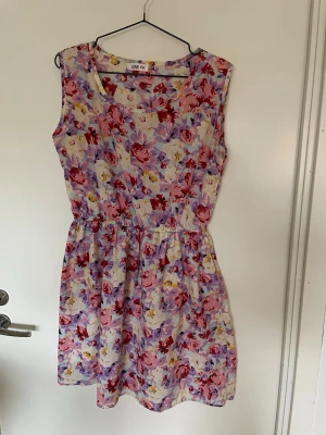 Blommig ärmlös klänning från LOVE FH - Söt ärmlös klänning med blommigt mönster i rosa, lila, rött och vitt. Klänningen har en rund halsringning och resår i midjan för en skön passform. Perfekt för sommarens varma dagar och ger en fräsch och färgglad look.