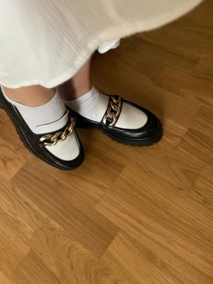 Chunky loafers med guldkedja - Säljer ett par svarta och vita chunky loafers med grov platt sula och rund tå. Skorna har en stor guldfärgad kedjedetalj framtill. 