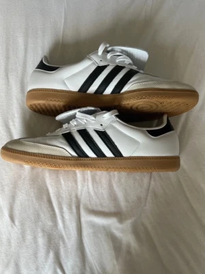 Adidas vita sneakers med gummisula - Säljer ett par klassiska Adidas sneakers i vitt läder med svarta ränder på sidorna och gummisula. Kostar 1250kr nypris och är knappast använda, storlek 48 2/3 men är aningen små i storleken så passar storlek 48 skulle jag säga!