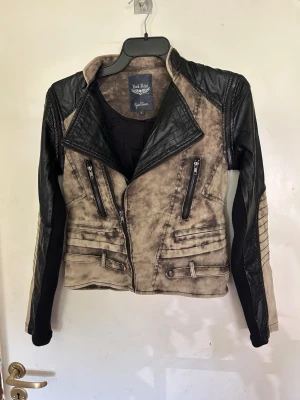 Rock Rebel jacka storlek S avtagbara ärmar - Snygg bikerinspirerad jacka från Rock Rebel i storlek S. Jackan har svart och beige tvättad look, dragkedjor framtill, quiltade detaljer på axlar och ärmar samt bältesdetaljer vid midjan. Perfekt för dig som gillar en edgy stil. Konstskinn och tyg. Avtagbara ärmar.