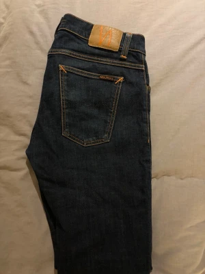 Mörkblå slim fit Nudie Jeans - Snygga mörkblå slim fit jeans från Nudie Jeans med klassiska fem fickor och kontrastsömmar. Jeansen har en smal passform och är tillverkade i denim med en lätt stretch för extra komfort. Perfekta för en trendig och avslappnad stil.
