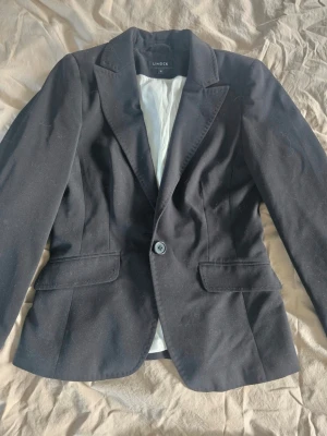 Svart kavaj från Lindex - Modellen är figursydd med axelvaddar, och upplevs som väldigt liten i storleken – är tajt på mig som vanligtvis bär storlek 34 eller 36. Fint skick med snygga detaljer.