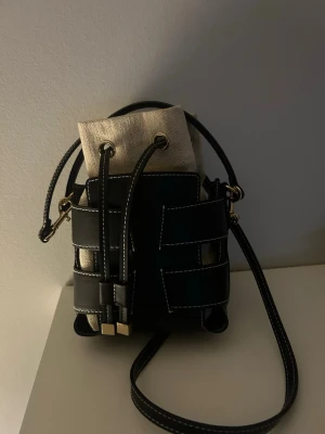 Svart och beige bucket bag - Trendig bucket bag i svart konstläder med vita kontrastsömmar och beige tyginsats. Väskan har justerbar axelrem och guldfärgade metalldetaljer. Stängs med dragsko och har en cool, modern design som sticker ut.