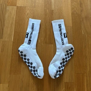 Vita fotbollsstrumpor med grepp - Säljer ett par vita fotbollsstrumpor med svarta grepp på undersidan och texten 'FOOTBALLSOCKS' längs skaftet. Strumporna är ribbade och har en sportig look, perfekta för dig som vill ha extra grepp i skorna.