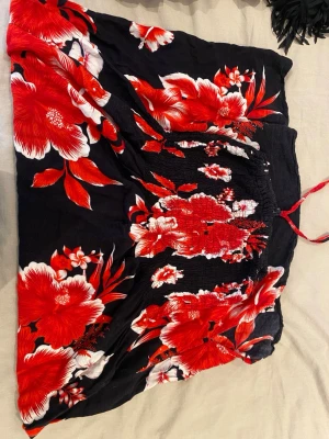 Svart kjol med röd blommig print - Supersnygg svart kjol med stora röda och vita blommor. Kjolen har smock i midjan och knytband för extra passform. Perfekt för sommarens alla äventyr och ger en färgstark vibe till din outfit.