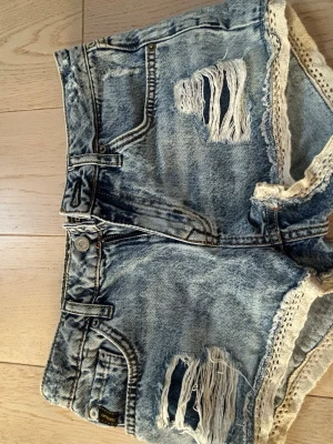 Blå denimshorts med spetsdetalj - Säljer ett par blå denimshorts med slitna detaljer och råa hål framtill. Shortsen har spetskant längs bensluten och klassisk femficksdesign, dom e från superdry och e i storlek 26 alltså S! Pris kan diskuteras, shortsens e low rise och bra skick! 