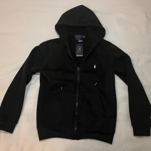Svart hoodie från Polo Ralph Lauren - Säljer en svart hoodie från Polo Ralph Lauren med dragkedja, huva och två fickor framtill. Klassisk vit logga broderad på bröstet och justerbara snören i huvan. Tillverkad i mjukt bomullsmaterial. Längd från axelhöjd till midjan: 60-62cm. Pris kan diskuteras😊