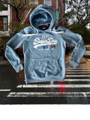 Ljusblå Hoodie från superdry - Snygg blå hoodie från Superdry med vit logga och japansk text på bröstet. Hoodien har en stor ficka framtill, dragsko i huvan och ribbade muddar vid ärmar och nederkant. Skit schysst hoodie som tyvärr blivit lite liten. Om du har frågar är det bara att fråga på. Pris går att diskutera med seriösa köpare. Kolla in min profil och se om du hittar något mer intressant. Mvh Larsen_Closet