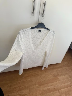 Vit spetsblus med v-ringning strl XL - Vit långärmad top i i spets med v-ringad hals. Toppens spetsmönster ger en romantisk känsla och den är perfekt för att lyfta en outfit. Snygg att styla med jeans eller kjol ny men saknar pristagg .