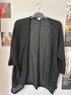 Svart öppen cardigan med mönster - Snygg svart cardigan med öppen front och diskret sicksackmönster. Lätt och luftig modell med trekvartsärmar, perfekt att slänga över en t-shirt eller topp. Materialet är tunt och genomskinligt vilket ger en avslappnad vibe.