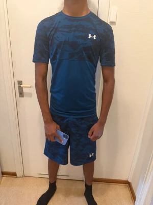 Blått träningsset från Under Armour - Säljer ett matchande träningsset från Under Armour i blått med mörkare kamouflagemönster. Setet består av en t-shirt med rund hals och korta ärmar samt shorts med elastisk midja. Perfekt för gymmet eller löpning. Skön outfit 