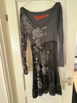 Grå blommig klänning Desigual - Unik grå klänning från Desigual med vita blommiga mönster och lång ärm. Klänningen har en snygg v-ringning och ett flowy fall som ger en cool och avslappnad vibe. Perfekt för dig som gillar statement-plagg med kreativa detaljer. Ett sidenband i grått saknas runt midjan.