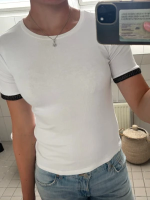 Vit t-shirt med svarta detaljer - Snygg vit t-shirt med rund halsringning och korta ärmar. Ärmsluten har svarta, glittriga detaljer som ger en cool kontrast. T-shirten är figurnära och gjord i mjukt, stretchigt material som känns skönt mot huden. Perfekt till jeans eller kjol. Den har liten fläck vid kragen. 